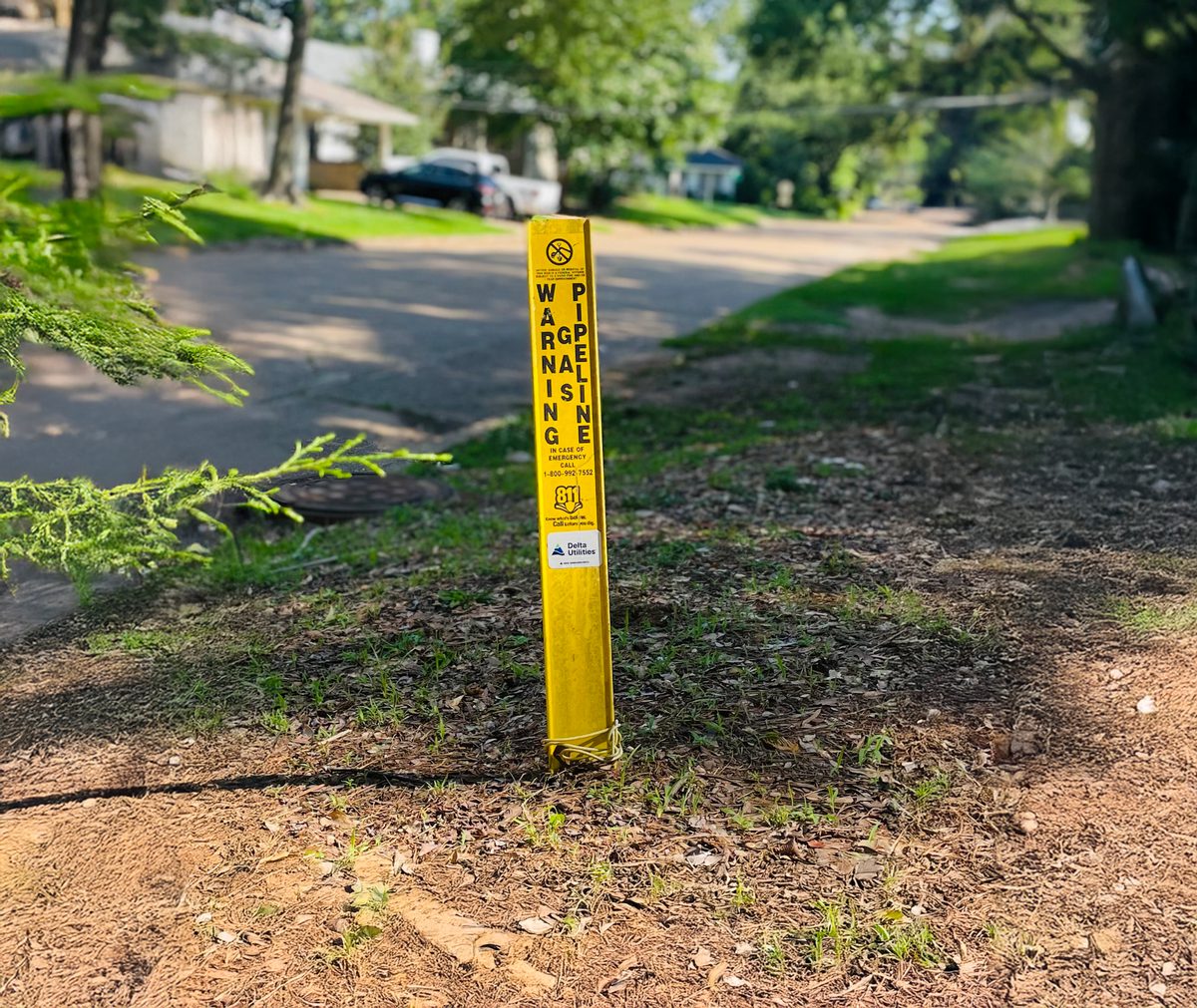 Pipeline Marker on a Natural Gas Pipe. There is a Delta Utilities logo at the top of the side of the pipe. On the left side is "CAUTION" in yellow letters with a black block background. "GAS PIPELINE" are on the right in black letters with a white background. Underneath those words, it reads, "LINE LOCATING TEST POINT". Underneath those words, it reads, "IN CASE OF EMERGENCY CALL 1-800-477-0177. Know what's below. Call before you dig."