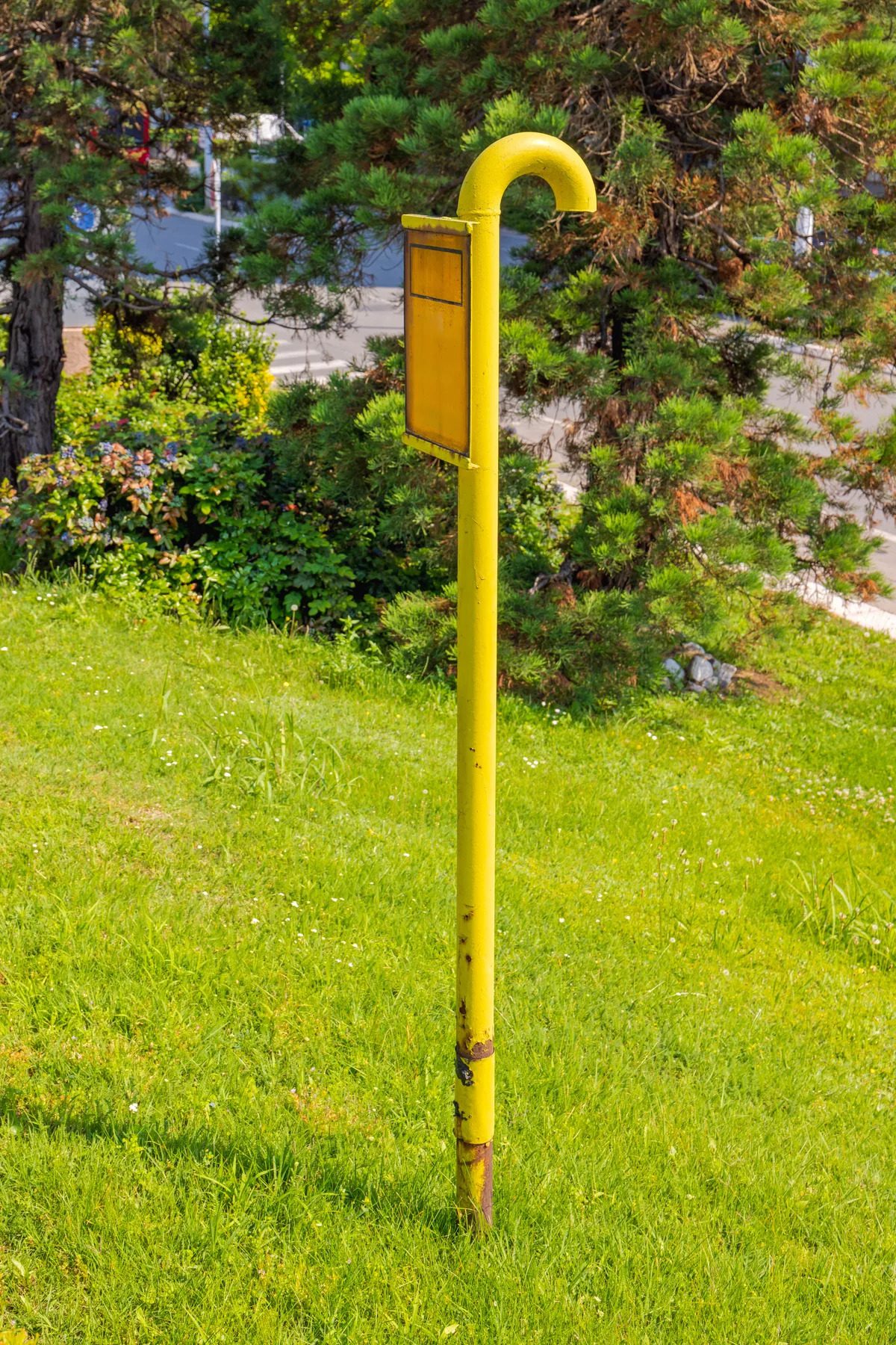 A yellow metal pipe with a curved top and a small rectangular sign is installed vertically in a grassy area near a road. The pipe appears weathered with some rust at the base, and it's surrounded by green grass and trees in the background.