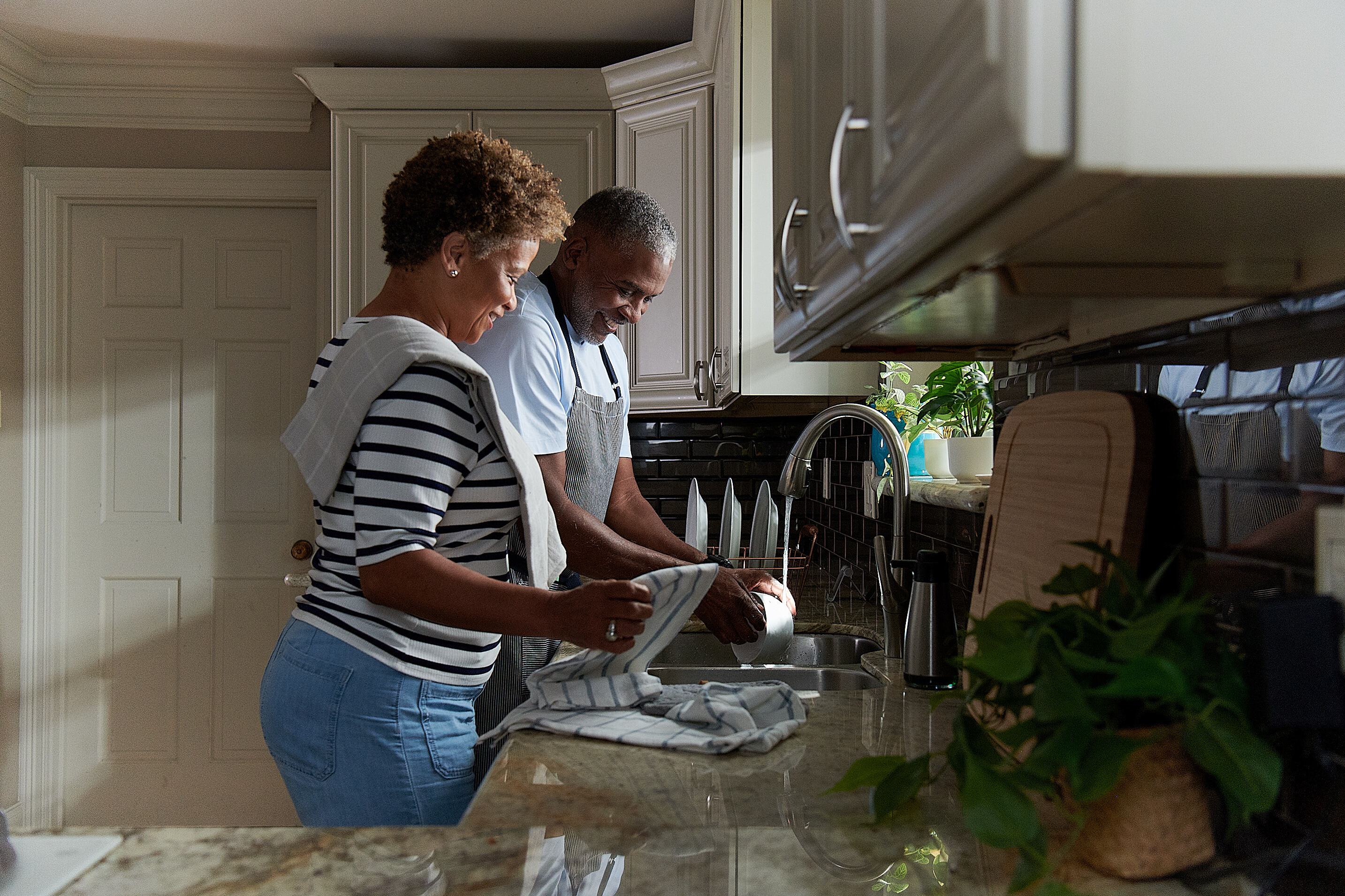 Older couple washing dishes with hot water from a natural gas powered sink in a home kitchen.